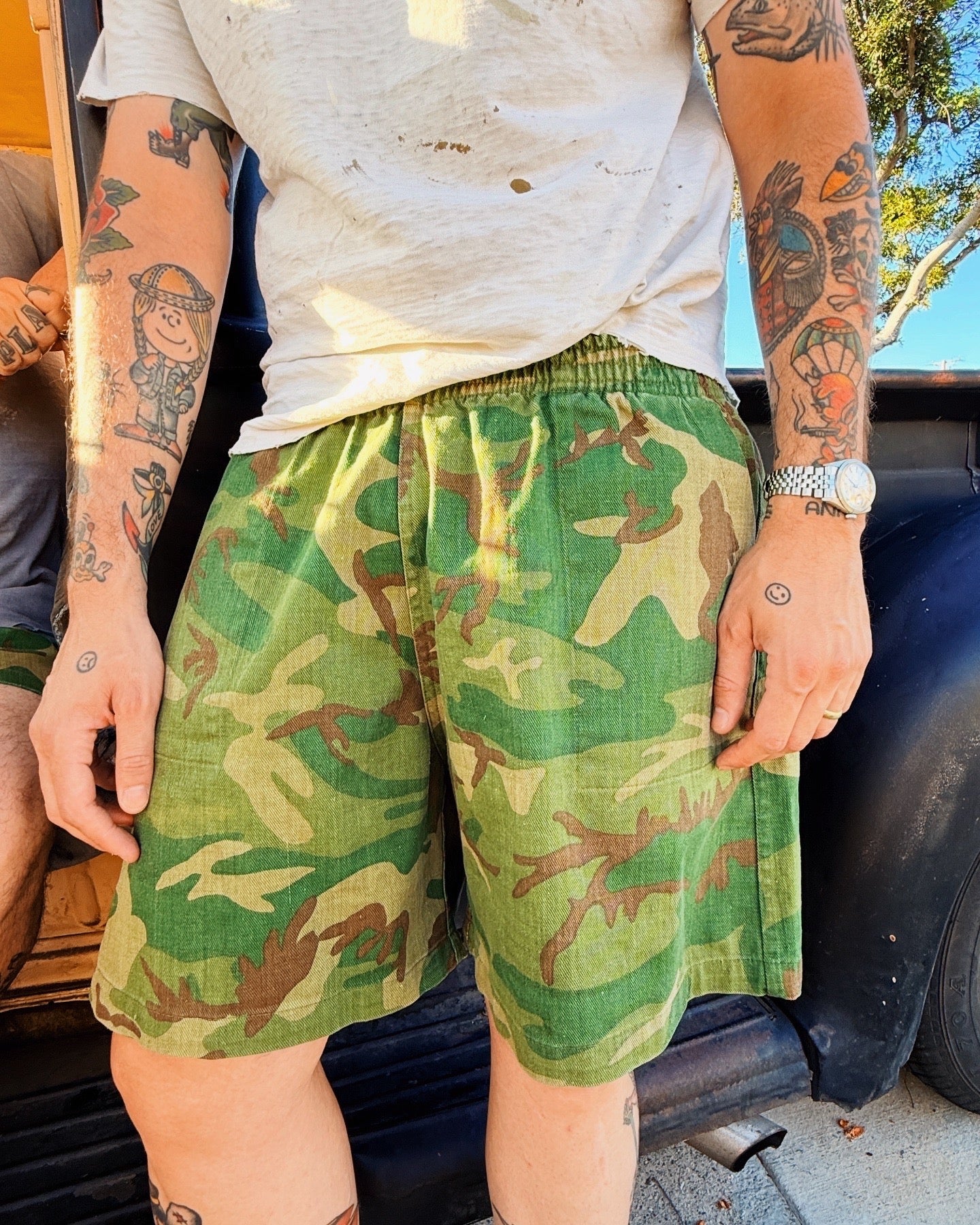 Person wearing green camouflage shorts with tattoos on arms, standing outdoors.