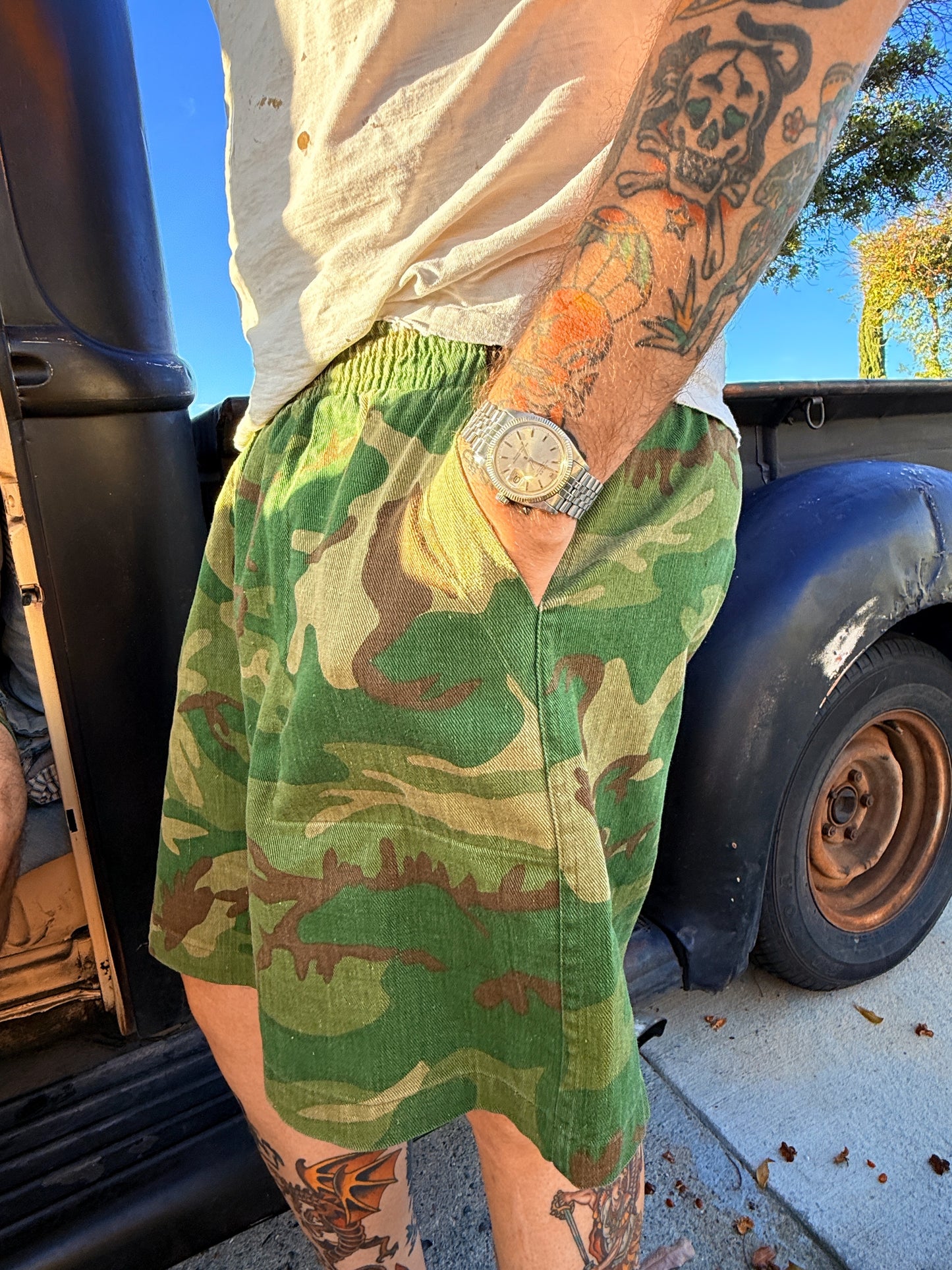 Person wearing camouflage shorts with a tattooed arm and watch, standing next to a vehicle.