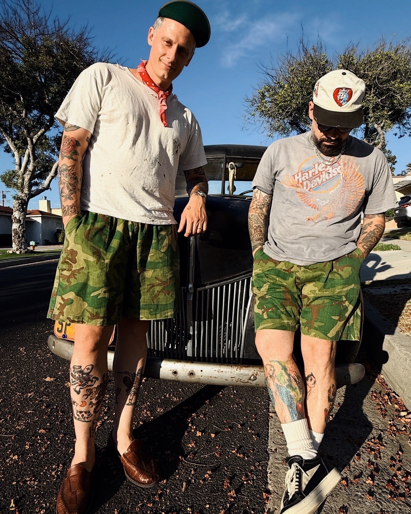 Two people wearing camouflage shorts standing in front of a vintage car.